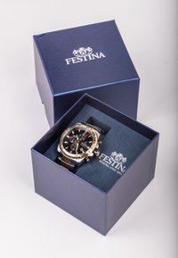 Auksinis ir sidabrinis nerūdijančio plieno laikrodis su juodu ciferblatu mėlyname dėkle. Dėklas turi minkštą mėlyną vidų ir "Festina" logotipą.