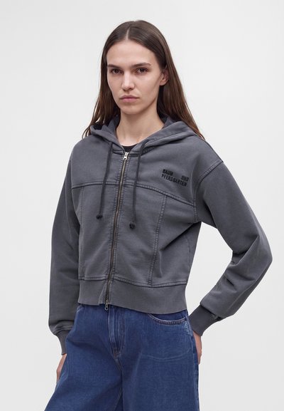 Femme aux longs cheveux portant un sweat à capuche gris zippé et un jean bleu, debout avec une main sur la hanche devant un fond uni.