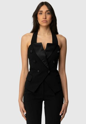 Donna che indossa un blazer nero senza maniche con collo halter e rever in raso, e pantaloni neri coordinati, in piedi davanti a uno sfondo semplice.