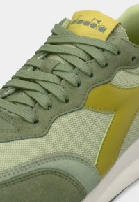 Világoszöld sportcipők hálós és velúr anyagok keverékével, texturált felülettel, kerek fűzőkkel és sárga logóval a nyelvén.