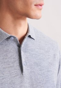 Nahaufnahme des unteren Gesichts und Halses eines Mannes, der ein hellgraues, geknöpftes Strickhemd mit zwei sichtbaren Knöpfen trägt.