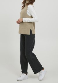 Beige neulottu hihaton poolopaita, jossa on ribbineulottu pinta ja sivuhalkiot, käytetty valkoisen pitkähihaisen paidan päällä, yhdistetty mustiin leveälahkeisiin housuihin ja valkoisiin lenkkareihin.