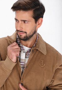 Brun corduroyjakke med krave, der har en broderet ankerdetalje. Underneath en ternet skjorte i hvide, lilla og blå farver.