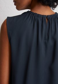 Oberer Rücken einer Frau in einem ärmellosen, dunkelblauen Blusentop mit gerafftem Ausschnitt und kleiner Schlüsselöffnung im Nacken.