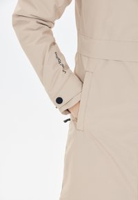 Beige vedenkestävä takki, jossa on sileä pinta, napitettavat hihansuut, sivutasku ja säädettävä naruvyötärö.