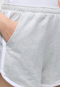Χέρι στην τσέπη ενός ανοιχτογκρι βαμβακερού σορτς με ελαστική μέση και λευκό περιθώριο, φορεμένο με λευκό τοπ.