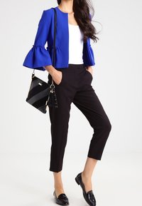 Veste courte bleue à manches évasées, associée à un pantalon noir ajusté et des mocassins noirs. Accessoire : un sac à main rayé avec pompon.