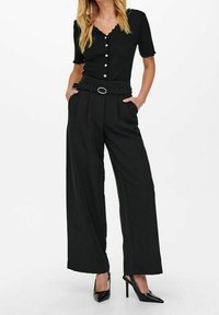 Black short-sleeve top with buttons and a scalloped hem, paired with high-waisted black wide-leg trousers and black high heels.