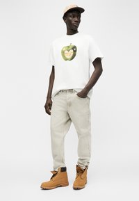 Άνδρας με λευκό μπλουζάκι με γραφικό πράσινο μήλο, ανοιχτό μπεζ παντελόνι, καφέ εργασιακά μποτάκια και μπεζ καπέλο μπέιζμπολ, στέκεται με το ένα χέρι στην τσέπη.