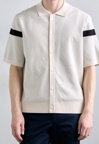 Polo a maniche corte in maglia color beige chiaro con motivo testurizzato, dotato di una patta bianca e di una striscia nera sulla spalla. Finitura liscia.