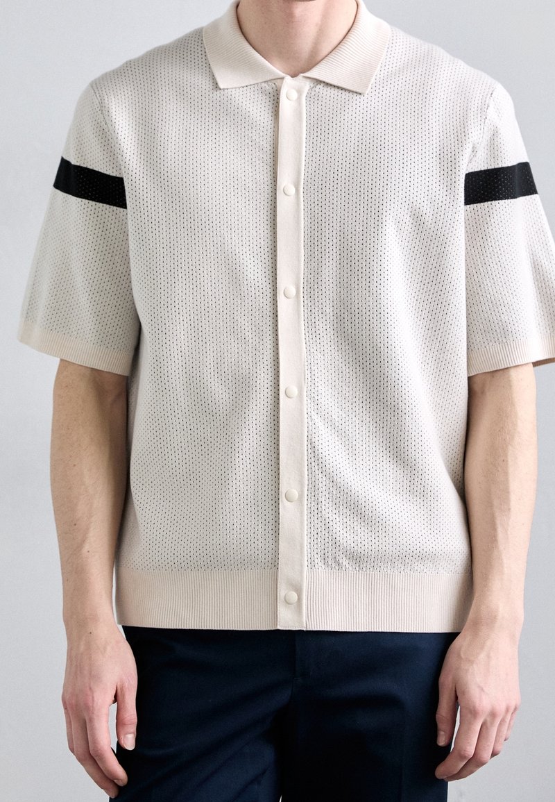 Polo a maniche corte in maglia color beige chiaro con motivo testurizzato, dotato di una patta bianca e di una striscia nera sulla spalla. Finitura liscia.