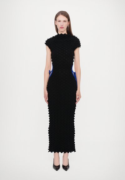 Robe noire avec des formes pyramidales texturées en 3D, encolure haute, manches courtes et liens accentués en bleu à la taille. Escarpins noirs portés.
