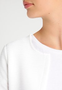 Weißer texturierter Stoffjacke mit einem runden Ausschnitt, getragen über einem schlichten weißen Hemd, das feine Rippungen und klare Linien zeigt.