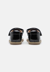 Zapatos infantiles de charol negro con puntera blanca, diseño redondeado, tira de velcro y suela de goma. La vista trasera muestra los detalles de costura.