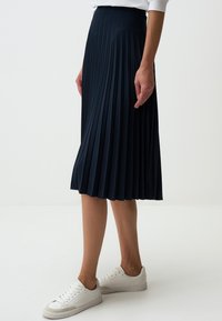 Marineblauwe geplooide rok van zachte stof, met een hoge tailleband en vloeiende silhouet. Draagt met witte sneakers.