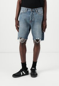Pantalones cortos de mezclilla azul claro con dobladillo deshilachado y detalles desgastados, combinados con calcetines negros y zapatillas negras con acentos blancos.