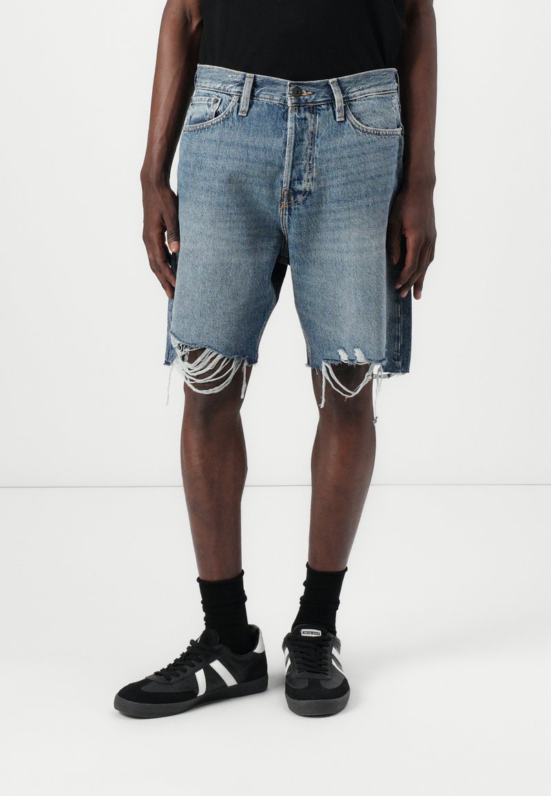 Pantalones cortos de mezclilla azul claro con dobladillo deshilachado y detalles desgastados, combinados con calcetines negros y zapatillas negras con acentos blancos.