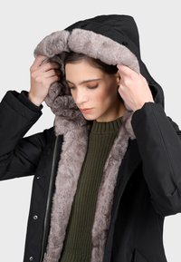 Černý zimní kabát s podšívkou z umělé kožešiny v světle šedé barvě. Kabát má zapínání na zip v přední části a pod ním je pletený svetr v olivově zelené barvě.