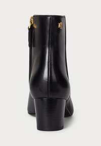 Black leather ankle boot with a rounded shape, mid-height block heel, rear zipper, and gold logo accent on the side. Smooth texture throughout.