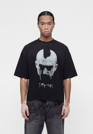 Giovane uomo con vitiligine che indossa una maglietta nera oversize con una grande grafica monocromatica del volto e jeans scuri con fantasia su uno sfondo semplice.