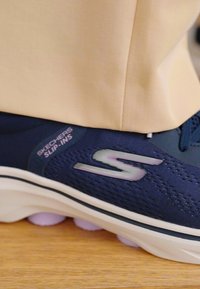 Marineblaue Skechers Slip-Ins verfügen über eine atmungsaktive, strukturierte Oberseite, eine glatte Sohle und ein reflektierendes Logo an der Seite, kombiniert mit einer hellbeigen Hose.