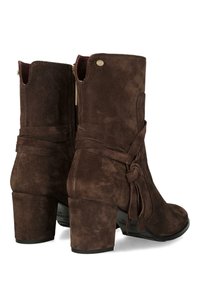 Botas de tobillo de ante marrón con un tacón grueso, que presentan un acento atado y un cierre de cremallera en la parte posterior. Superficie texturada y diseño elegante.