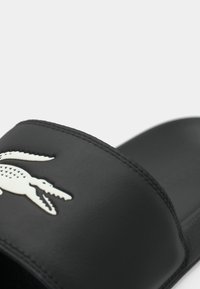 Sandale à glissière noire avec une tige synthétique lisse, présentant un logo crocodile blanc et des détails de couture. Semelle intérieure rembourrée, design à talon ouvert.