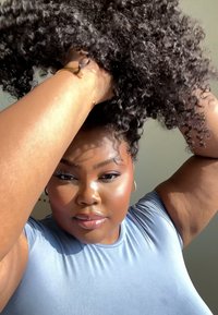 Modell med krøllete svart hår, iført en lyseblå tettsittende topp. Har polert hud, store hoop-øreringer, og drar hår opp med den ene hånden.