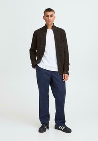 Dunkelbraune, gerippte Zip-Jacke über einem weißen T-Shirt, kombiniert mit navyblauen Hosen und schwarzen Sneakers mit weißen Streifen.