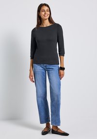 Frau steht da und trägt ein schwarzes Oberteil mit drei Viertelärmeln, blaue gerade Jeans, schwarze Ballerinas und ein schwarzes Handgelenkaccessoire, während sie leicht lächelt.