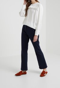 Blusa bianca a maniche lunghe con dettagli a volant, abbinata a pantaloni eleganti blu navy e scarpe slip-on in suede arancione con un accento hardware decorativo.