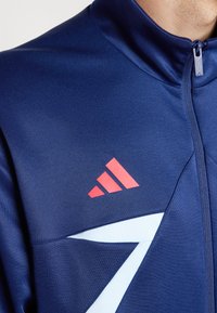 Námornícky modrá zipová bunda s vysokým golierom, svetlomodrými akcentmi a červenými vertikálnymi pruhmi s logom Adidas na ľavej hrudi.