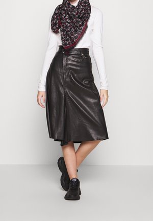 Femme portant une jupe midi en cuir noire, un haut blanc à manches longues, une écharpe noire à motifs et des baskets noires, debout les jambes croisées.