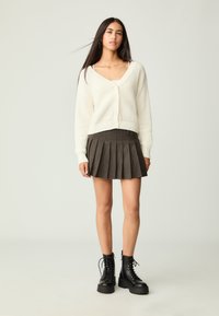 Weißer gerippter Strickcardigan mit V-Ausschnitt und Knopfverschluss, kombiniert mit einem braunen plissierten Rock. Schwarze Schnürplattformstiefel runden den Look ab.