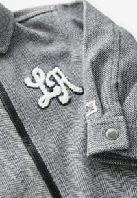 Veste gris chevron avec fermeture éclair noire, écusson blanc texturé « LA », petite étiquette logo et bouton-pression en métal au poignet.