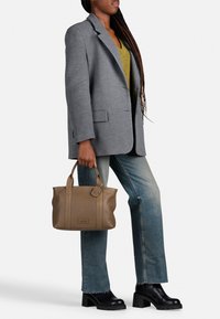 Grijze wollen blazer over een groene top, blauwe jeans en zwarte plateau boots. Houdt een tan leren schoudertas vast met een label.