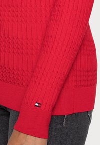 Red knit sweater with textured cable design and ribbed cuffs. Features a small logo patch in blue, white, and red on the sleeve.