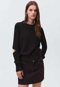 Črna pletena jopica z rebrastimi manšeti in robom, ki ima izrezane detajle na komolcih, kombinirana s črno denim krilom z žepi.