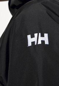 Chaqueta impermeable negra con textura suave, que presenta un logotipo bordado en blanco "HH" en el área del hombro.