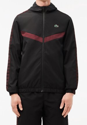 Homme portant une veste noire zippée avec une bande en chevron bordeaux sur la poitrine et des bandes assorties à motifs bordeaux sur les manches.
