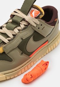 Zöld bőr és hálós sneaker bézs és narancssárga kiegészítőkkel, textúrázott fűzőkkel és egy plusz feltekert narancssárga fűzővel.