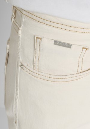 Close-up van crèmekleurige spijkerbroek met een voorvak met contrasterende stiksels en een metalen merkembleem met de tekst "ANTONY MORATO."