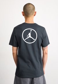 Camiseta de algodón gris oscuro con un gran logo blanco de Air Jordan en la parte posterior. Cuello redondo clásico y mangas cortas.