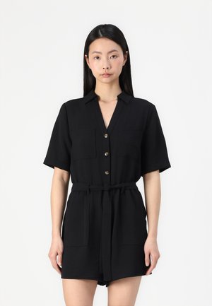 Combinaison noire à manches courtes avec un col, des boutons sur le devant, deux poches poitrine et une ceinture nouée à la taille. Tissu texturé.