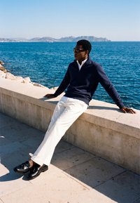 Homme portant des lunettes de soleil, un pull bleu marine et un pantalon blanc, assis sur un mur de mer avec de l'eau bleue et des montagnes au loin en arrière-plan.