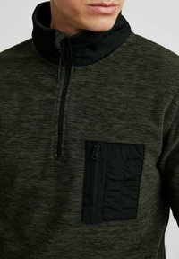 Groene fleece pullover met een zwarte hoge kraag en een ritszak. Gelaagd materiaal met een contrasterend glad paneel op de borst.