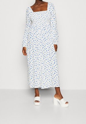 Femme portant une robe blanche longueur cheville avec imprimé floral bleu, manches longues bouffantes, encolure carrée, et talons compensés blancs ouverts à l'avant.