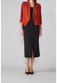 Chaqueta corta roja sobre un vestido negro ajustado con una abertura hasta la rodilla. Tejido suave, corte relajado y escote barco. Tacones altos negros.