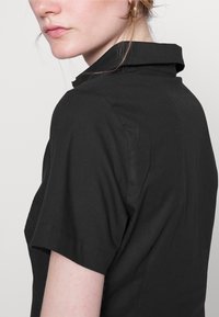 Camicetta nera a maniche corte con colletto a punta e una texture simile al cotone. Il design include una silhouette pulita con cuciture sottili.