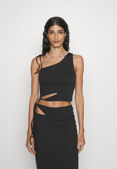 Femme portant un crop top noir à une épaule et une jupe assortie avec des découpes, se tenant devant un fond blanc uni.